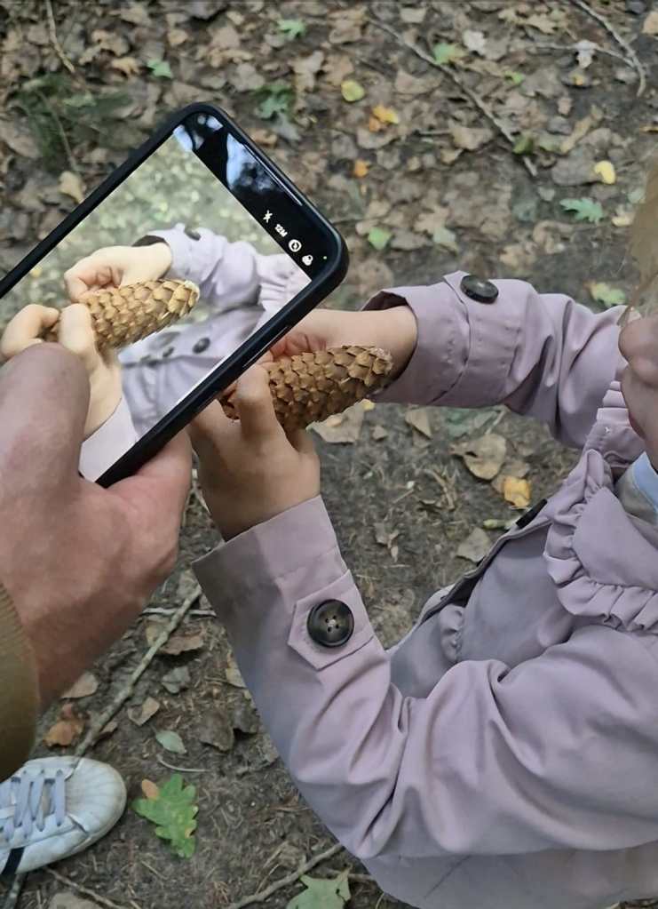 Een kind houd een dennenappel vast. Een volwassene maakt een foto van het gevonden voorwerp met de telefoon.